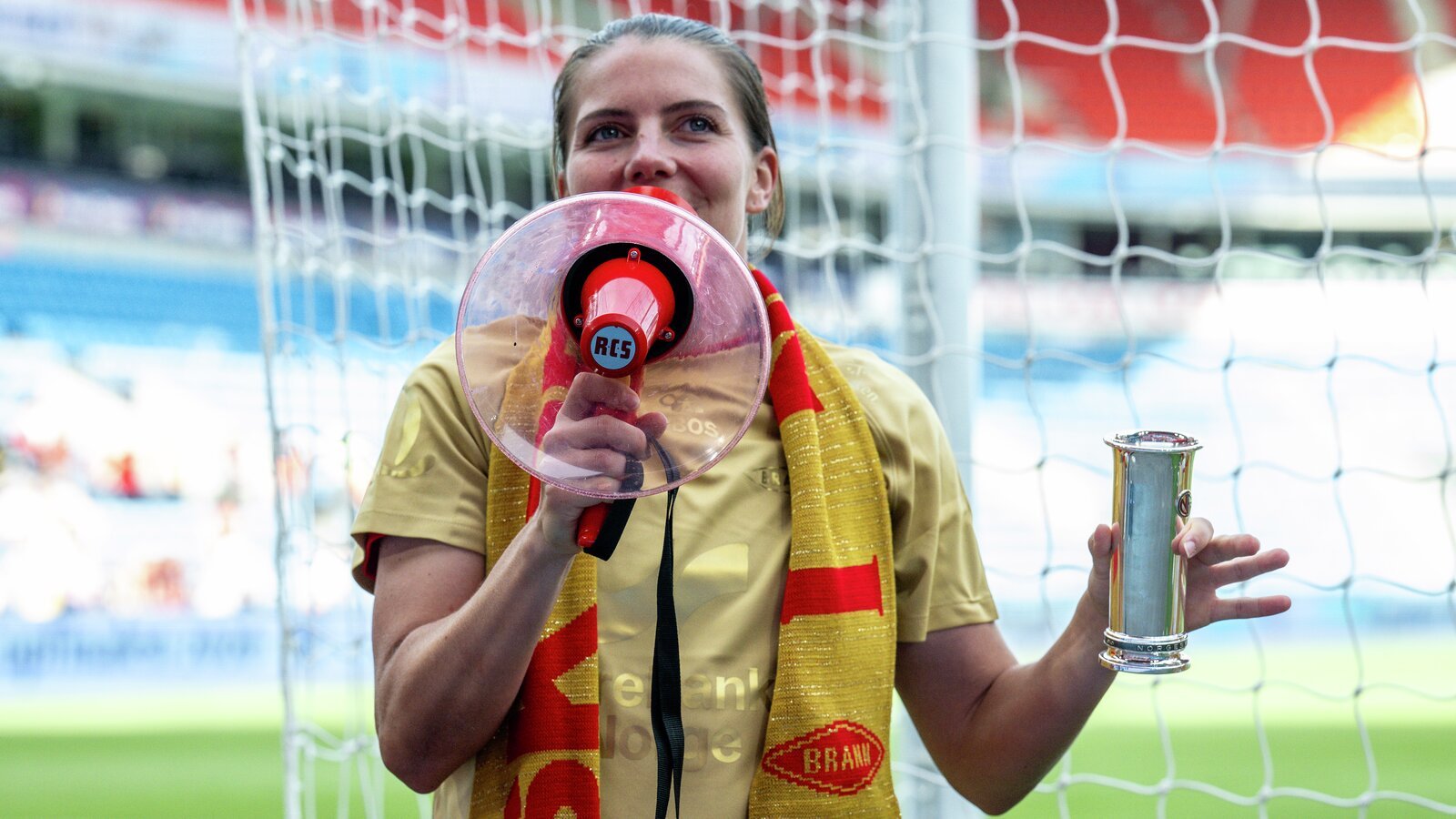 En kvinne i en gulltrøye holder en rosa megafon og et sølvtrofé i nærheten av et fotballmål.